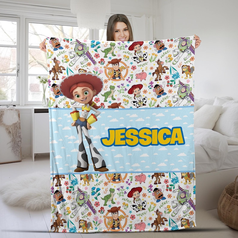 May include: A white fleece blanket featuring a repeating pattern of Toy Story characters including Jessie, Woody, Buzz Lightyear, Rex, and Mr. Potato Head, with a central light blue horizontal band displaying the name JESSICA in yellow text.