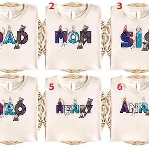 Puede incluir: Seis camisetas color crema con diferentes nombres impresos en una fuente colorida y estilizada. Los nombres incluyen "DAD", "MOM", "SIS", "BRO", "HENRY" y "ANNA". Cada camiseta presenta personajes de dibujos animados.