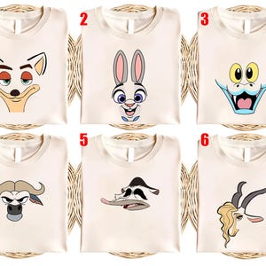 May include: Six cream-colored t-shirts, each featuring a different cartoon character face. The designs include a fox, a rabbit, a dragon, a buffalo, a duck, and a gazelle. The shirts are displayed on a woven surface.