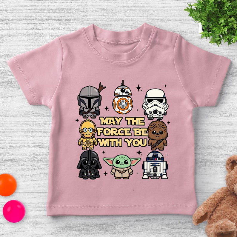May include: Pink short-sleeved t-shirt featuring cartoon illustrations of Star Wars characters, including The Mandalorian, BB-8, and Darth Vader. The phrase "May the Force Be With You" is printed in the center of the shirt.