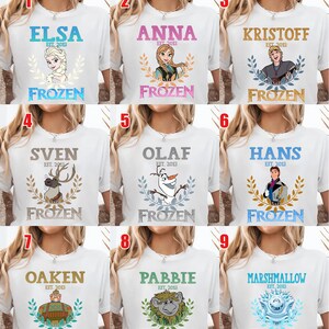 Puede incluir: Camisetas blancas con personajes de la pel&iacute;cula "Frozen". Cada camiseta tiene el nombre de un personaje, el texto "EST. 2013" y la palabra "FROZEN" en una fuente decorativa. Los personajes incluyen a Elsa, Anna, Kristoff, Sven, Olaf, Hans, Oaken, Pabbie y Marshmallow.