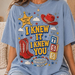 May include: A light blue t-shirt featuring a graphic print with a yellow sheriff star labeled INFINITY AND BEYOND, a red cowboy hat, red cowboy boots, three stacked alphabet blocks with T, S, 5, and a beaded necklace with YOU'VE GOT A FRIEND IN ME.