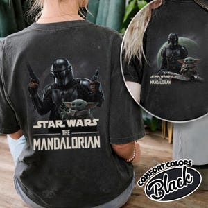 Puede incluir: Camiseta negra con un dise&ntilde;o de Star Wars que presenta a The Mandalorian y Grogu. El Mandaloriano se muestra con armadura, sosteniendo blasters, con Grogu debajo. El texto "STAR WARS THE MANDALORIAN" est&aacute; impreso en blanco.