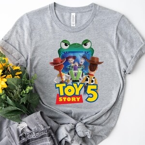 May include: A heather gray t-shirt featuring a graphic print of characters from Toy Story, including Woody, Jessie, and Buzz Lightyear, positioned in front of a green frog-shaped screen. The text Toy Story 5 is printed in blue and yellow.