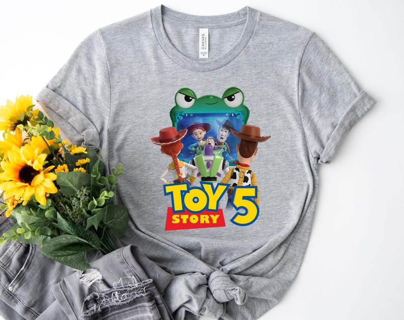 May include: A heather gray t-shirt featuring a graphic print of characters from Toy Story, including Woody, Jessie, and Buzz Lightyear, positioned in front of a green frog-shaped screen. The text Toy Story 5 is printed in blue and yellow.