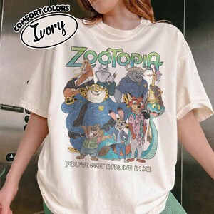 May include: Ivory-colored t-shirt featuring a colorful graphic of characters from the animated film "Zootopia." The text "Zootopia" is in green, and the phrase "You've Got a Friend in Me" is below the characters. The shirt has short sleeves.