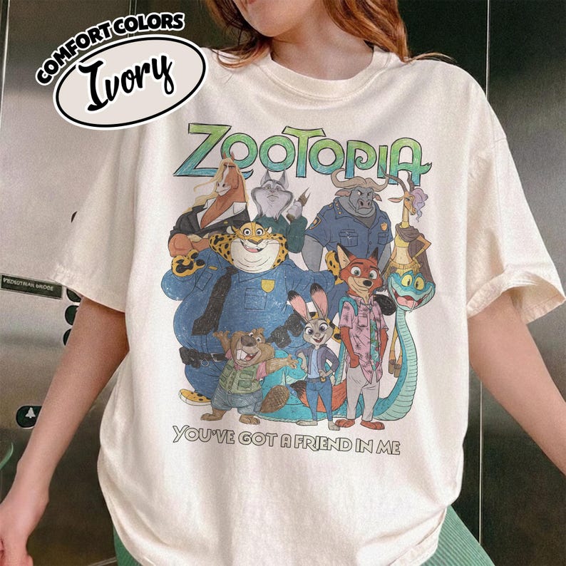 May include: Ivory-colored t-shirt featuring a colorful graphic of characters from the animated film "Zootopia." The text "Zootopia" is in green, and the phrase "You've Got a Friend in Me" is below the characters. The shirt has short sleeves.