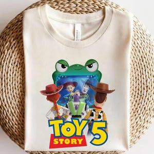 May include: A cream-colored t-shirt featuring a graphic of characters from Toy Story looking at a green frog-shaped tablet screen. The text "TOY STORY 5" is printed in large blue and yellow letters below the characters on the front of the shirt.