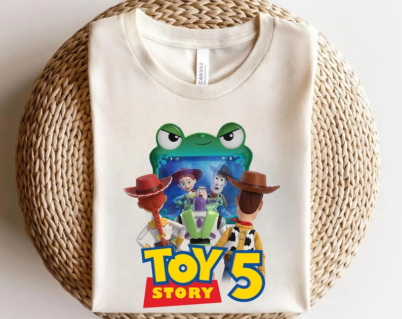 May include: A cream-colored t-shirt featuring a graphic of characters from Toy Story looking at a green frog-shaped tablet screen. The text "TOY STORY 5" is printed in large blue and yellow letters below the characters on the front of the shirt.