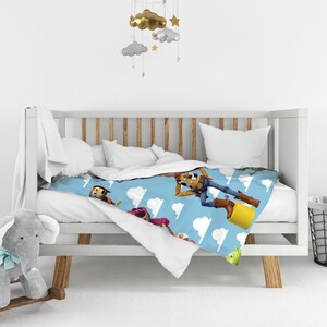 K&ouml;nnte beinhalten: Eine hellblaue Decke mit einem sich wiederholenden Muster aus Toy Story-Figuren wie Woody, Lotso und Slinky Dog, durchsetzt mit wei&szlig;en Wolkenformen. Die Decke liegt &uuml;ber einem wei&szlig;en Holzbettchen, daneben steht ein grauer Pl&uuml;schelefant.