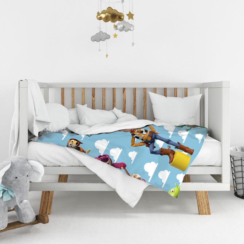 K&ouml;nnte beinhalten: Eine hellblaue Decke mit einem sich wiederholenden Muster aus Toy Story-Figuren wie Woody, Lotso und Slinky Dog, durchsetzt mit wei&szlig;en Wolkenformen. Die Decke liegt &uuml;ber einem wei&szlig;en Holzbettchen, daneben steht ein grauer Pl&uuml;schelefant.