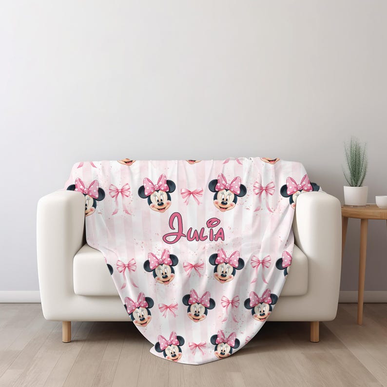 May include: A soft throw blanket featuring a repeating pattern of Minnie Mouse faces and pink bows on a light pink and white vertical striped background. The name Julia is printed in a cursive pink font in the center of the blanket.