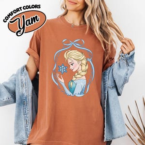 May include: Rust-colored t-shirt featuring a cartoon illustration of a woman with blonde hair and a blue dress, holding a snowflake. The illustration is framed by a blue ribbon design. The words "COMFORT COLORS" and "Yam" are printed on the top left.