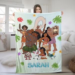 K&ouml;nnte beinhalten: Eine hellblaue Fleecedecke mit einem farbenfrohen Grafikdruck der Vaiana-Figuren, darunter Maui, Vaiana, Pua, Heihei und Kakamora. Das Design zeigt tropische Bl&auml;tter und Blumen sowie den Namen SARAH in gro&szlig;en blauen Buchstaben am unteren Rand.