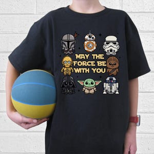 May include: Black t-shirt featuring Star Wars characters including Darth Vader, Yoda, and Chewbacca. The phrase "MAY THE FORCE BE WITH YOU" is printed in the center. The shirt is worn by a person holding a blue and yellow ball.
