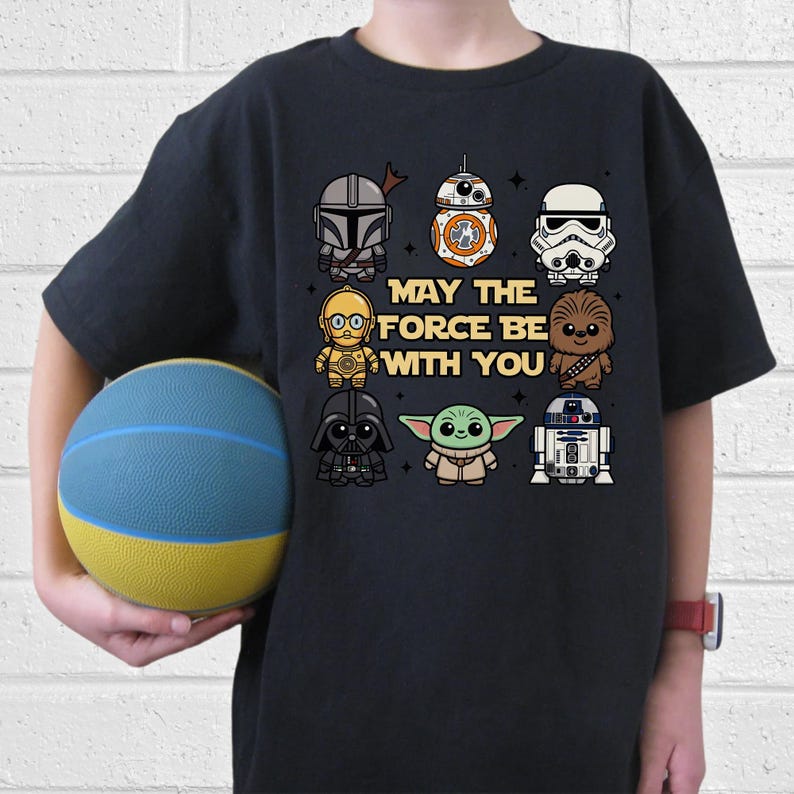 May include: Black t-shirt featuring Star Wars characters including Darth Vader, Yoda, and Chewbacca. The phrase "MAY THE FORCE BE WITH YOU" is printed in the center. The shirt is worn by a person holding a blue and yellow ball.
