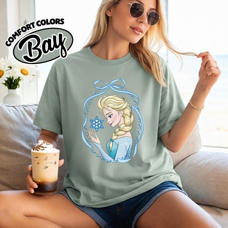 May include: Sage green t-shirt featuring a cartoon illustration of a blonde woman with a braid, wearing a blue dress, holding a snowflake. The design is framed by a blue ribbon and lace-like border. The text "COMFORT COLORS Bay" is at the top.
