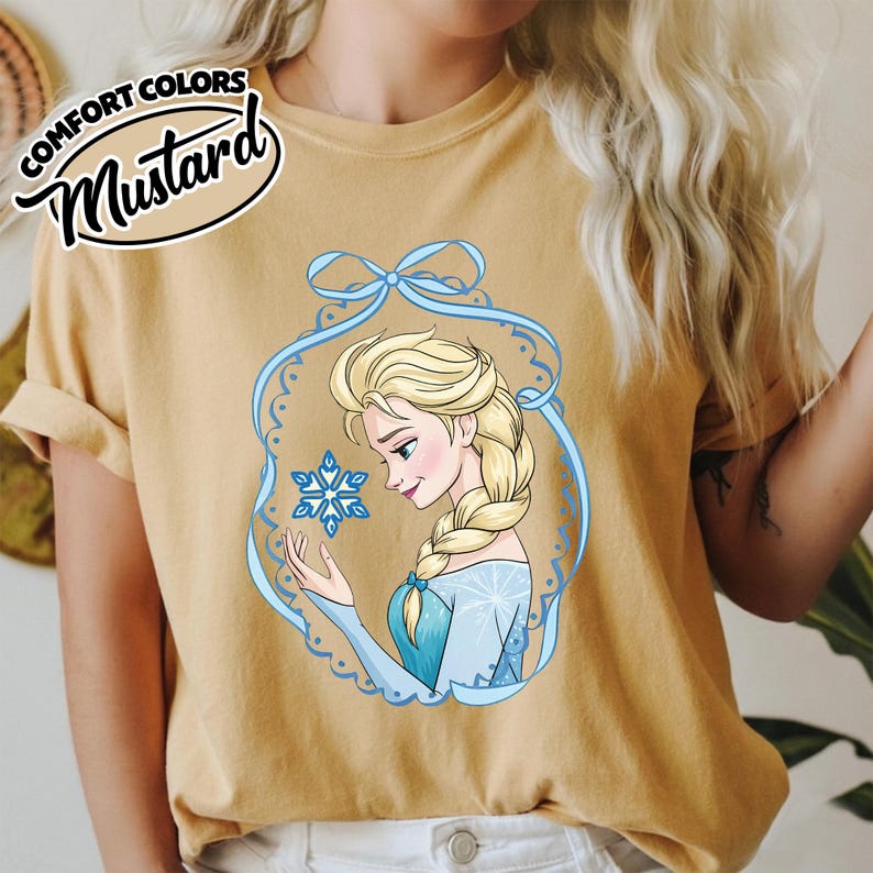 May include: Mustard-colored t-shirt featuring a cartoon illustration of a blonde woman with a braid, wearing a blue dress and holding a snowflake. The illustration is framed by a blue decorative border. The text "COMFORT COLORS" and "Mustard" are in the upper left corner.