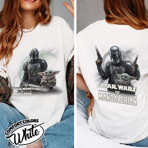 Puede incluir: Camiseta blanca con un gr&aacute;fico de The Mandalorian y Grogu de Star Wars. La parte delantera muestra a los personajes en un veh&iacute;culo, la trasera los muestra con blasters. La camiseta incluye el texto "STAR WARS THE MANDALORIAN".