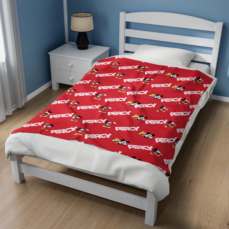 Puede incluir: Una manta de forro polar roja con un patr&oacute;n repetitivo de ilustraciones de Mickey Mouse y el nombre Percy impreso en letras may&uacute;sculas blancas. La manta est&aacute; sobre una estructura de cama blanca con una almohada y un colch&oacute;n blancos.