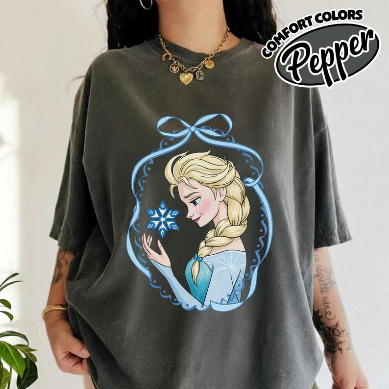 May include: Dark gray t-shirt featuring a cartoon illustration of a blonde woman in a blue dress holding a snowflake. The design is framed by a blue decorative border. The text "COMFORT COLORS Pepper" is printed on the upper right.