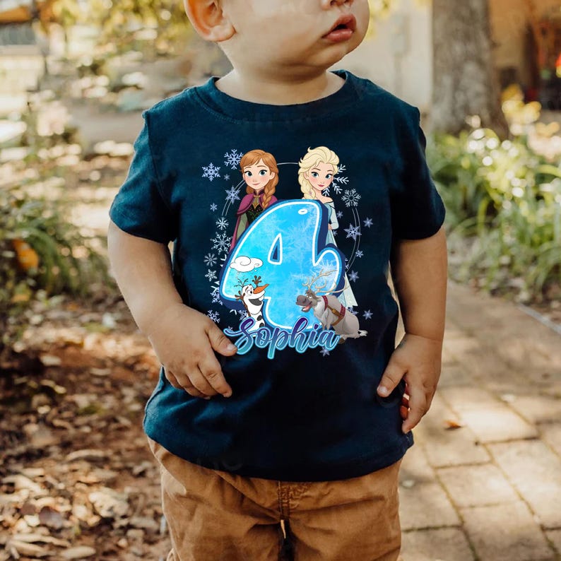 Pode incluir: Camiseta azul marinho para crian&ccedil;as pequenas com um gr&aacute;fico colorido do n&uacute;mero 4 e os nomes dos personagens Anna e Elsa do filme Frozen. O nome "Sophia" est&aacute; escrito abaixo. A camiseta &eacute; decorada com flocos de neve.