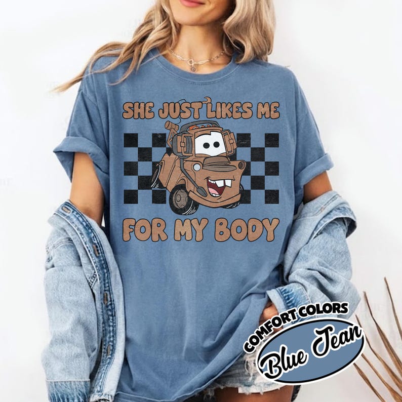 May include: A blue short-sleeve t-shirt featuring a graphic of a brown tow truck character with a headset. The design includes a black and white checkered background and the text "SHE JUST LIKES ME FOR MY BODY" in brown letters. The shirt has a crew neckline.