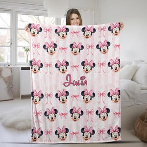 May include: A soft fleece blanket featuring a repeating pattern of Minnie Mouse faces with pink bows and small pink bows on a light pink and white vertical striped background. The name Julia is printed in a cursive pink font in the center.