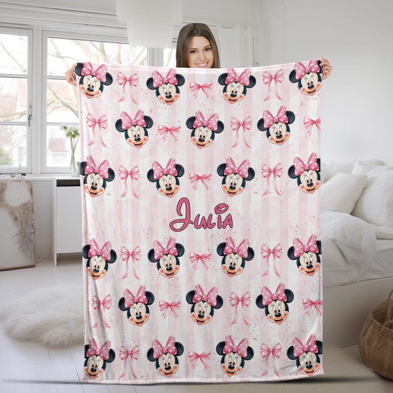 May include: A soft fleece blanket featuring a repeating pattern of Minnie Mouse faces with pink bows and small pink bows on a light pink and white vertical striped background. The name Julia is printed in a cursive pink font in the center.