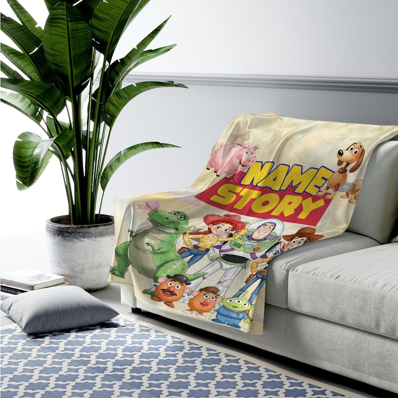 May include: A cream-colored throw blanket featuring a colorful graphic print of Toy Story characters including Buzz Lightyear, Woody, Jessie, Rex, Hamm, Slinky Dog, Mr. Potato Head, and an alien, with the text NAME STORY printed in bold yellow letters.