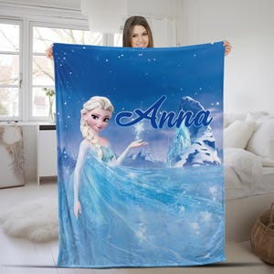 May include: A rectangular blue fleece blanket featuring a printed graphic of a blonde character in a light blue dress with a braid, standing near an icy mountain landscape. The name Anna is printed in a large, dark blue script font across the top.