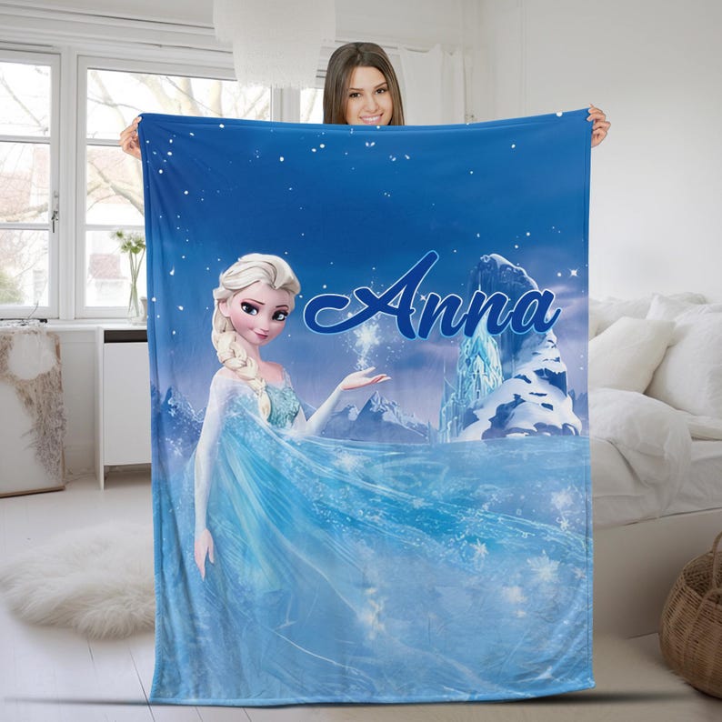 May include: A rectangular blue fleece blanket featuring a printed graphic of a blonde character in a light blue dress with a braid, standing near an icy mountain landscape. The name Anna is printed in a large, dark blue script font across the top.