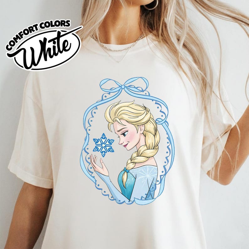 May include: White Comfort Colors t-shirt featuring a cartoon illustration of a blonde woman in a blue dress holding a snowflake. The illustration is framed by a blue decorative border. The shirt has the words "Comfort Colors White" printed on the top left.