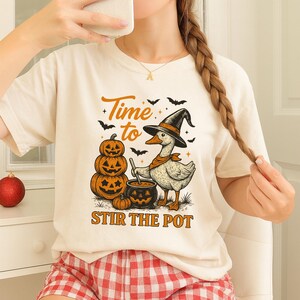May include: Cream-colored t-shirt with a Halloween-themed graphic. The design features a goose wearing a witch hat and stirring a cauldron, with pumpkins and bats. Text reads "Time to Stir the Pot."