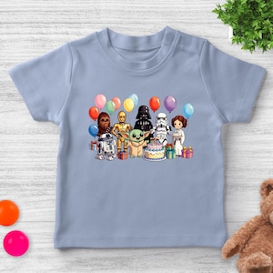 K&ouml;nnte beinhalten: Hellblaues T-Shirt mit einer farbenfrohen Grafik von Star Wars-Charakteren, darunter R2-D2, Chewbacca, C-3PO, Darth Vader, ein Sturmtruppler, Prinzessin Leia und Baby Yoda, die sich um eine Geburtstagstorte mit Luftballons versammelt haben.