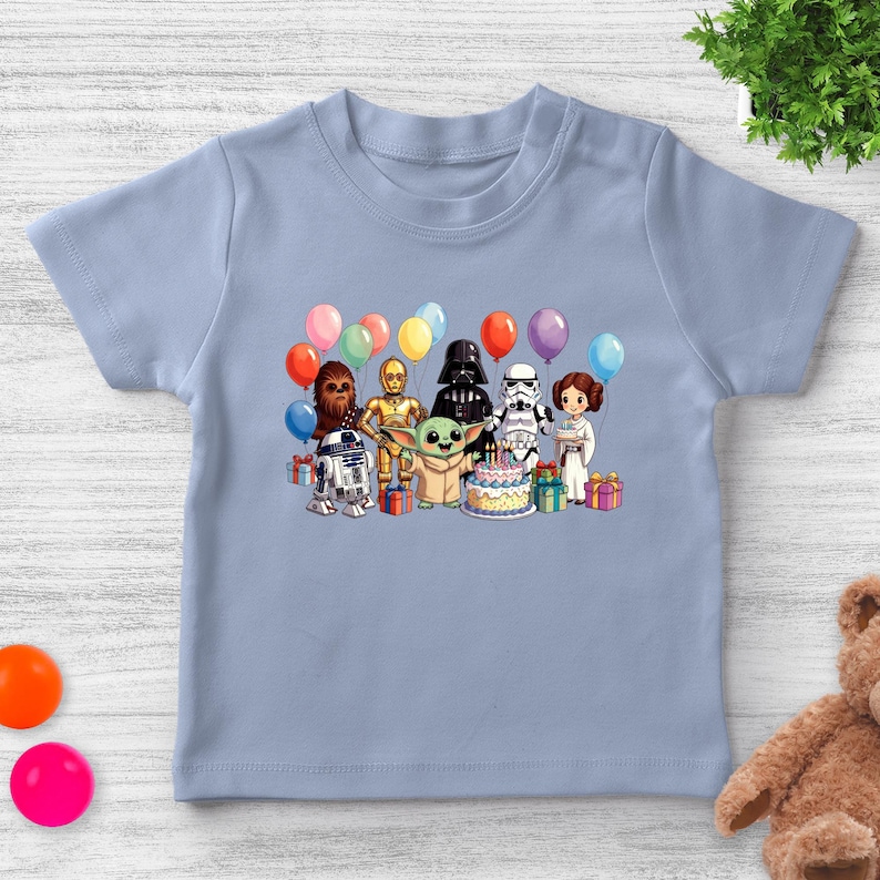 K&ouml;nnte beinhalten: Hellblaues T-Shirt mit einer farbenfrohen Grafik von Star Wars-Charakteren, darunter R2-D2, Chewbacca, C-3PO, Darth Vader, ein Sturmtruppler, Prinzessin Leia und Baby Yoda, die sich um eine Geburtstagstorte mit Luftballons versammelt haben.