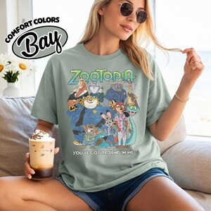 May include: Sage green t-shirt featuring a colorful graphic of characters from the animated film "Zootopia." The text "Zootopia" is above the graphic, and "You've Got a Friend in Me" is below. The shirt also has the Comfort Colors Bay logo.