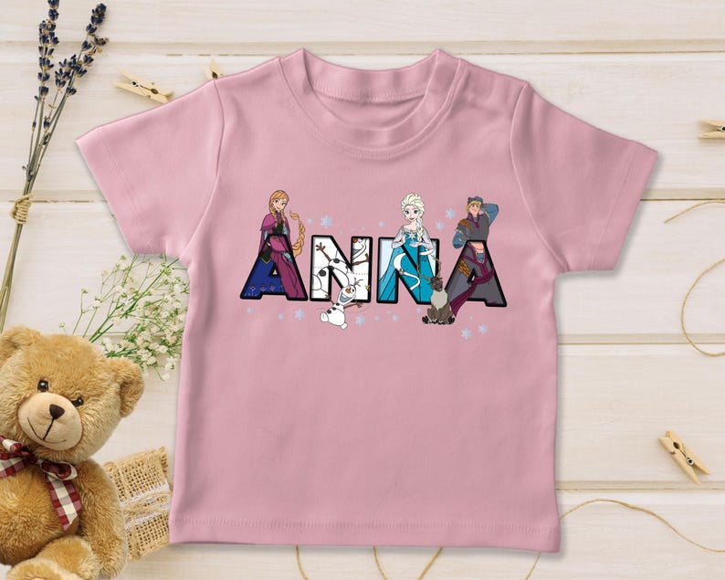 Puede incluir: Una camiseta rosa claro con el nombre "ANNA" en letras grandes y coloridas. Cada letra est&aacute; dise&ntilde;ada con un personaje diferente de la pel&iacute;cula, con copos de nieve y estrellas alrededor de las letras. La camiseta es de material suave.