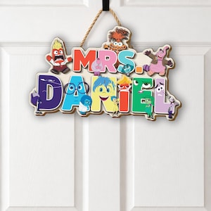 May include: A decorative wooden wall sign featuring the text MRS. DANIEL in large, colorful, block-style letters. The sign is adorned with various animated character illustrations from the movie Inside Out, including Joy, Sadness, and Anger.