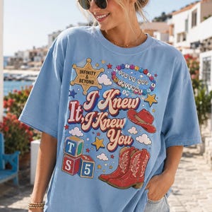 K&ouml;nnte beinhalten: Ein hellblaues T-Shirt mit kurzen &Auml;rmeln und einem Grafikdruck mit dem Text "I Knew It, I Knew You", einem Sheriffstern "Infinity & Beyond", einer Perlenkette, einem roten Cowboyhut, roten Cowboystiefeln und bunten Spielw&uuml;rfeln.