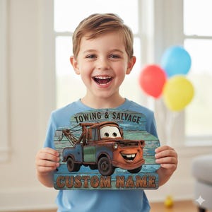 Puede incluir: Un letrero de madera con el personaje Mater de la pel&iacute;cula Cars, con el texto "TOWING & SALVAGE" y "CUSTOM NAME". El letrero tiene un aspecto r&uacute;stico y desgastado con una combinaci&oacute;n de colores azul y marr&oacute;n. La imagen la sostiene un ni&ntilde;o sonriente.