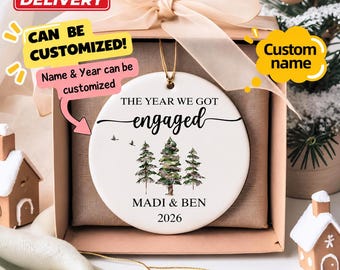 May include: A round white ceramic ornament featuring the text THE YEAR WE GOT engaged, a watercolor illustration of three pine trees, two small birds, and the names MADI & BEN with the year 2026 printed in black ink, suspended by a gold string.