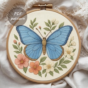 Puede incluir: Diseño de mariposa bordada en un aro de madera. La mariposa tiene alas azules con bordes marrones, rodeada de flores melocotón, naranjas y blancas con hojas verdes. El texto "PDF pattern" está en la esquina superior izquierda.