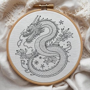 K&ouml;nnte beinhalten: Schwarz-wei&szlig; gesticktes Drachendesign auf wei&szlig;em Stoff, gespannt in einem h&ouml;lzernen Stickrahmen. Der Drache ist detailliert mit Schuppen und Krallen. Der Hintergrund ist ein weicher, wei&szlig;er Stoff. Der Rahmen hat einen Durchmesser von ca. 20 cm.