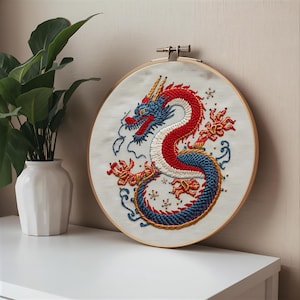 K&ouml;nnte beinhalten: Gesticktes Drachenkunstwerk in einem Holzreifen. Der Drache hat rote, wei&szlig;e und blaue F&auml;den mit aufwendigen Details. Das Kunstwerk wird auf wei&szlig;em Hintergrund mit einer Pflanze in einer wei&szlig;en Vase links gezeigt.