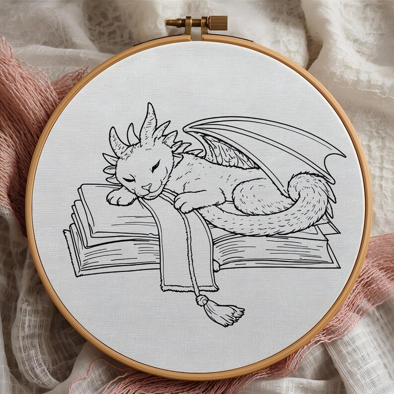 Puede incluir: Un bastidor de bordado en blanco y negro con un drag&oacute;n durmiendo sobre libros. El drag&oacute;n tiene alas y cola. El bordado est&aacute; sobre tela blanca, en un aro de madera.