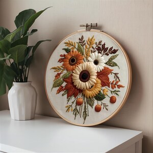 May include: A round embroidery hoop featuring a floral design with sunflowers and autumn leaves in shades of orange, yellow, cream, and green. The embroidery is set against a cream-colored fabric, and the hoop is made of wood.