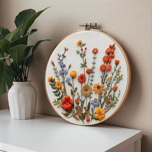 K&ouml;nnte beinhalten: Ein farbenfroher Stickrahmen mit Blumenmuster. Das Design zeigt gestickte Blumen in Orange-, Gelb-, Blau- und Rott&ouml;nen auf wei&szlig;em Stoff. Der Holzrahmen wird auf einer wei&szlig;en Oberfl&auml;che pr&auml;sentiert. Ein handgefertigtes Kunstwerk.