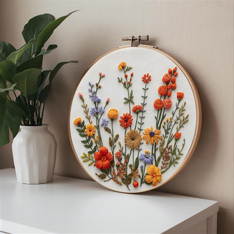 K&ouml;nnte beinhalten: Ein farbenfroher Stickrahmen mit Blumenmuster. Das Design zeigt gestickte Blumen in Orange-, Gelb-, Blau- und Rott&ouml;nen auf wei&szlig;em Stoff. Der Holzrahmen wird auf einer wei&szlig;en Oberfl&auml;che pr&auml;sentiert. Ein handgefertigtes Kunstwerk.