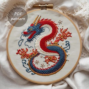K&ouml;nnte beinhalten: Gesticktes Drachendesign in einem Holzreifen. Der Drache hat rote, wei&szlig;e und blaue F&auml;den mit goldenen Akzenten. Der Text "PDF pattern" befindet sich in einem Kreis oben links. Die Stickerei befindet sich auf einem hellen Stoff.